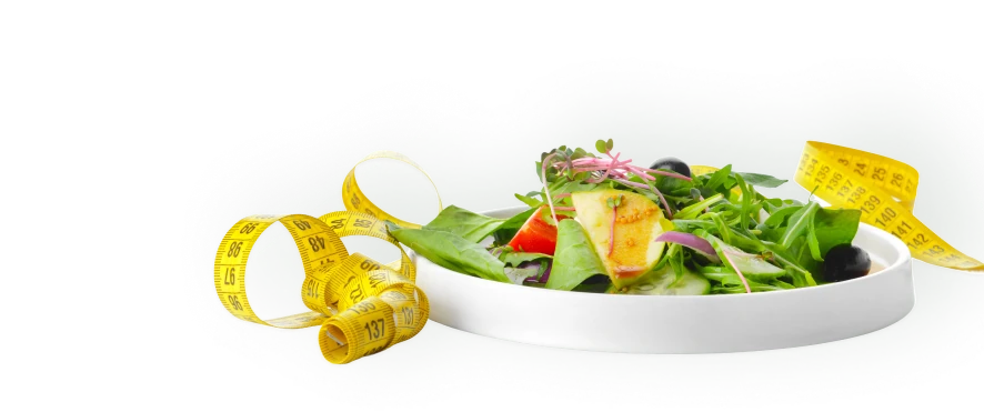Biała miska z kolorową sałatką z liści szpinaku, rukoli, plastrów ogórka, kawałków pomidora i czarnych oliwek. Obok leży żółta miarka krawiecka, symbolizująca dietę i odchudzanie.